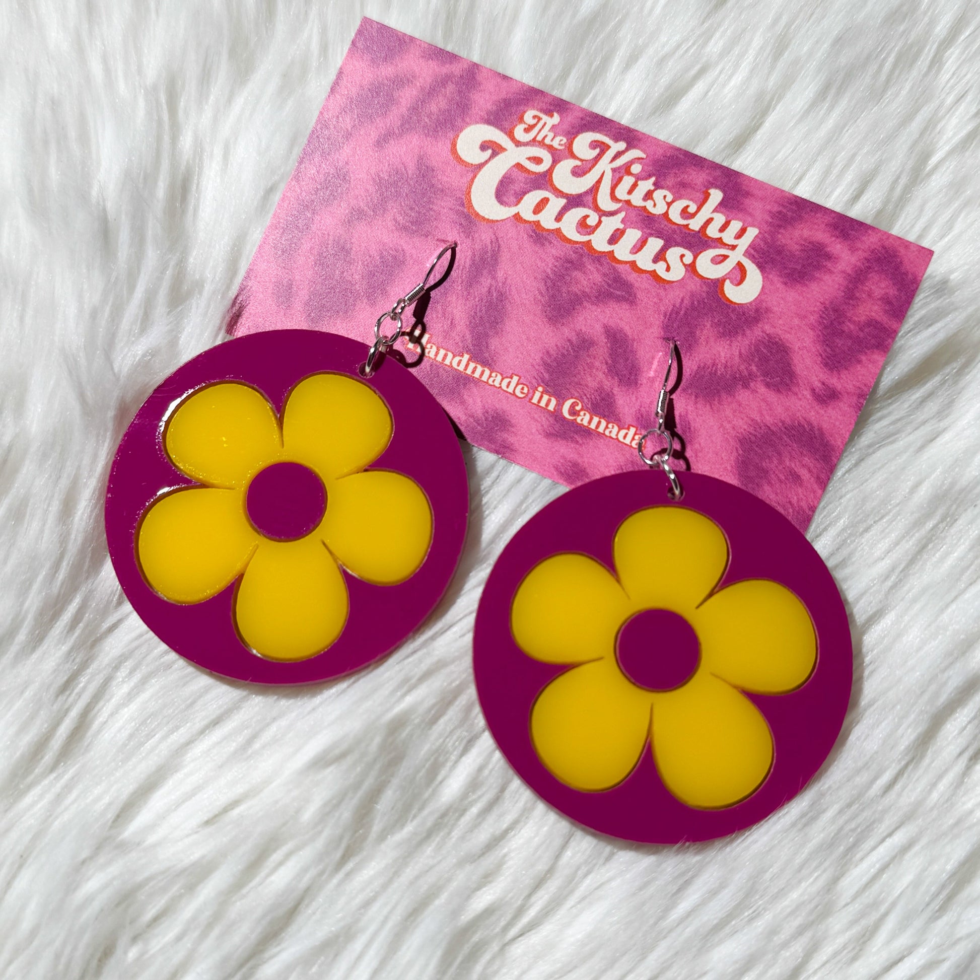 Flower-shaped earrings with purple and yellow design on a pink card with 'Kitschy Cactus' branding, placed on a white furry surface.